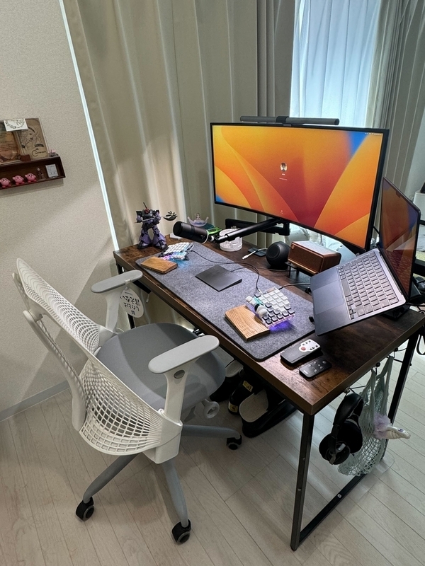 1個前の写真にさらに物が加わり、机環境が充実している。大きなモニターとアーム付きのマイクが導入されている。キーボードも割れた形式のものに進化している。