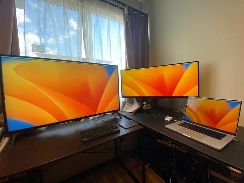 長机に特大サイズのモニターが2台、直角に配置されている。手前にはMacのノートPCが置かれている。特大デスクは昇降式。