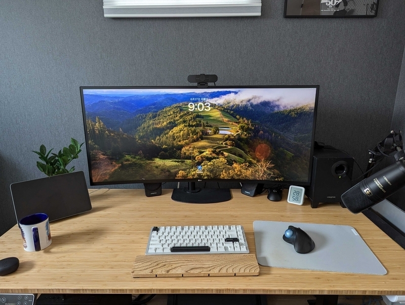 明るい色の木材を使った大きな机に、大きなモニターとキーボード、木目調のアームレストが置かれている。右側にマウスパッドとマイスがあり、左奥には小さなモニターもある。