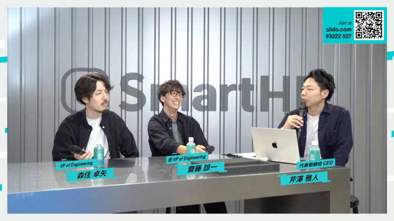 配信画面のキャプチャ：大きな銀テーブルに登壇者3名がかけている。テロップで名前が表示されており、左から森住さん、斎藤さん、芹澤さん。背景はSmartHRのロゴが書かれたグレーのソリッドな壁。