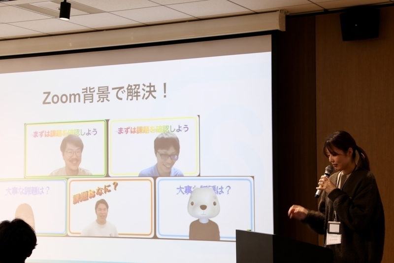 演台に立ち登壇中のeminemさん。スクリーンに投影された資料には「課題 is 何？」と書かれた程よくださいZoom背景を設定した写真が5枚貼られている