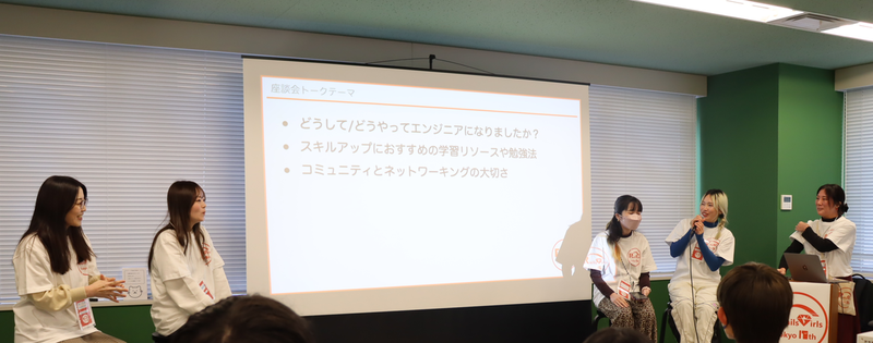 女性が５名スクリーンの前にいる。スクリーンには座談会トークテーマ「どうして/どうやってエンジニアになりましたか？」「スキルアップにおすすめの学習リソースや勉強法」「コミュニティとネットワーキングの大切さ」と書かれたスライドが映されている。