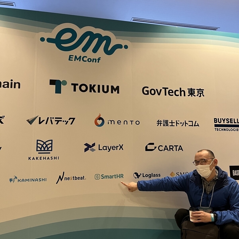男性がEMConf JP 2025のスポンサーが掲載されているパネルの前でSmartHRのロゴを指さしている