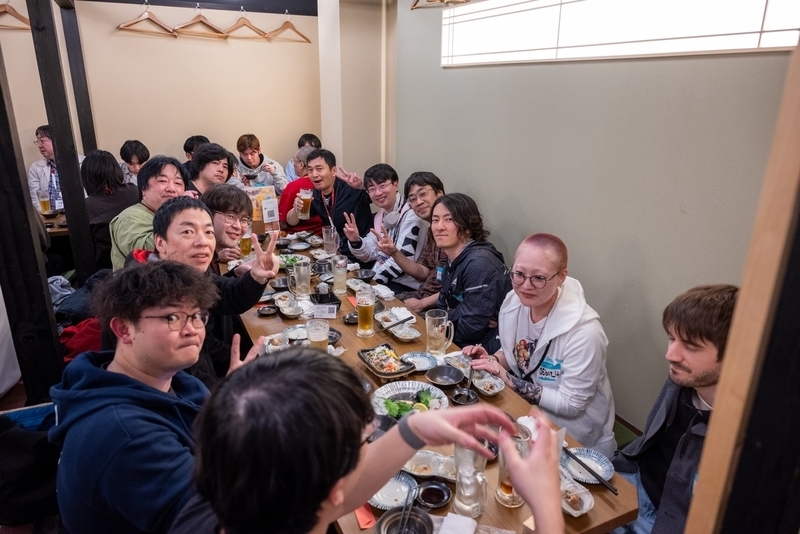 12名の参加者が料理とお酒を囲ってピースサインをしている。