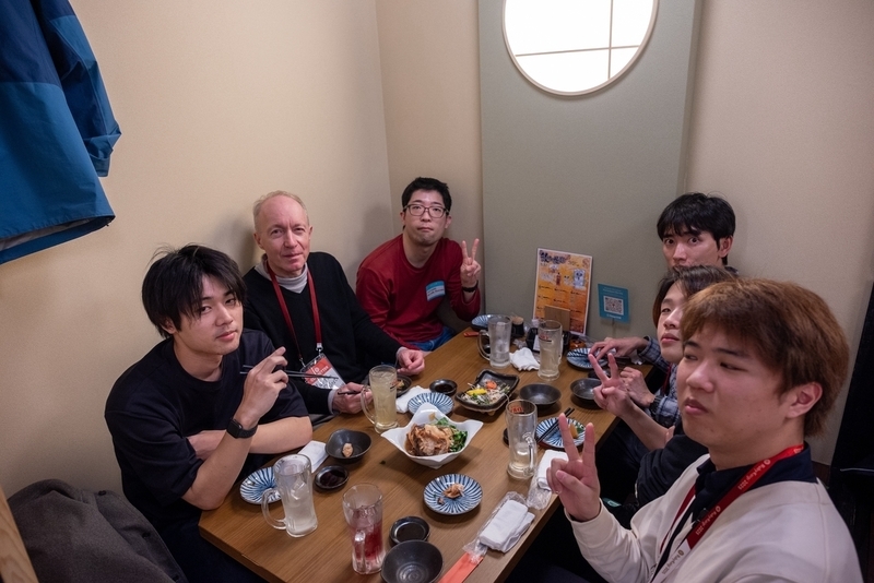 6名の参加者がお酒と食事を囲みながら写真に対してポーズをしている。
