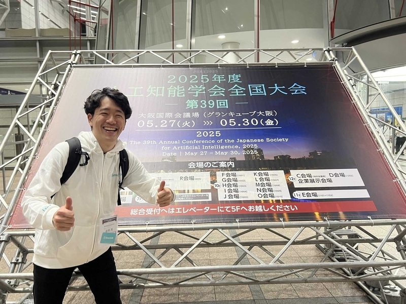 人工知能学会の看板の前で登壇者が両手をグッドをしながら満面の笑顔の写真