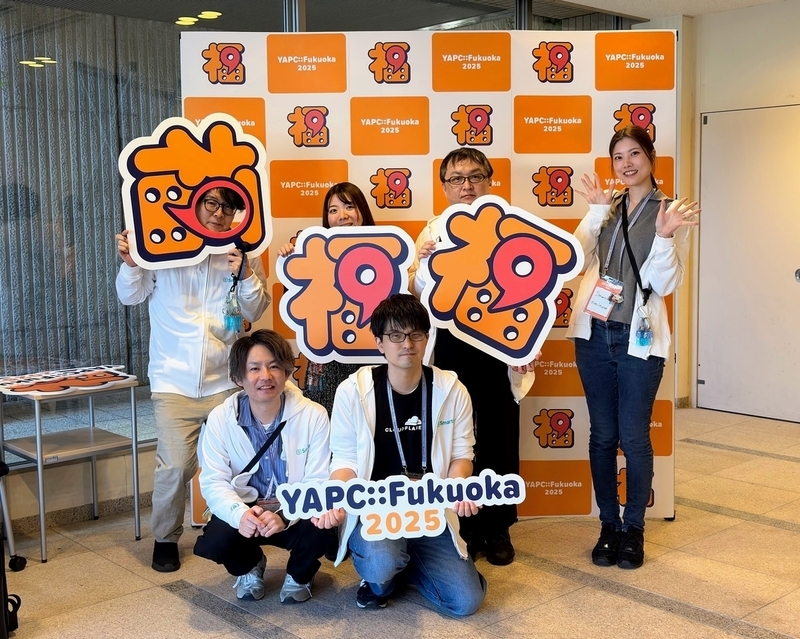 YAPC::Fukuoka 2025のフォトブースパネルの前で、SmartHRパーカーを着た6人がYAPC::Fukuoka 2025のロゴなどを持っている