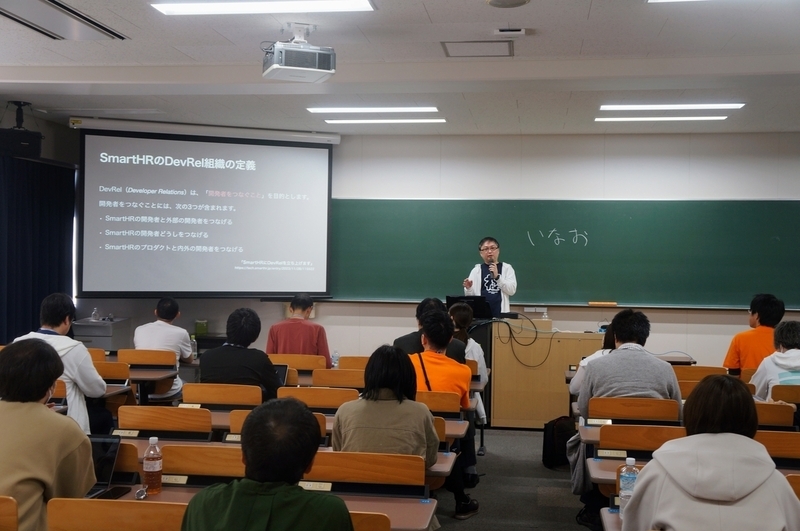 教壇で登壇している。スクリーンにはスライドが映され、黒板には「いなお」と書かれている