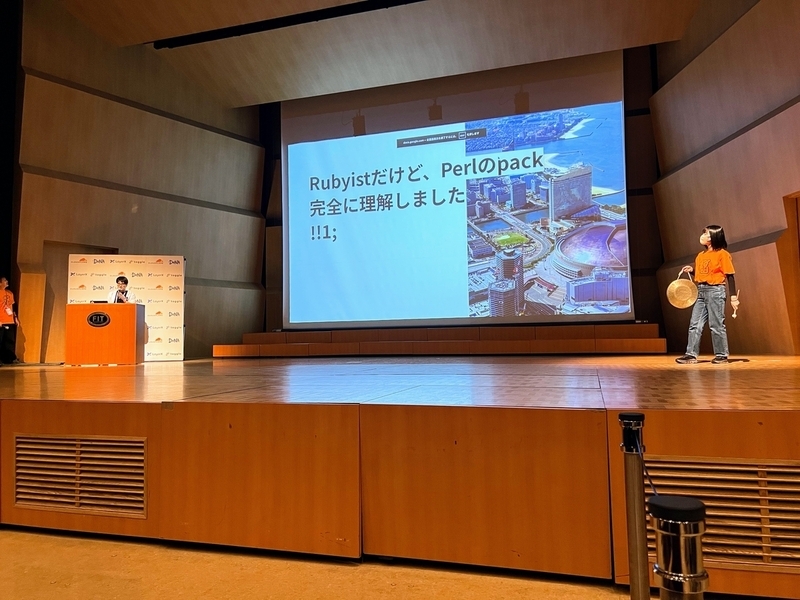 メインステージの中央に巨大スクリーンがあり、向かって左側で登壇している。右側にはドラを持った人が立っている