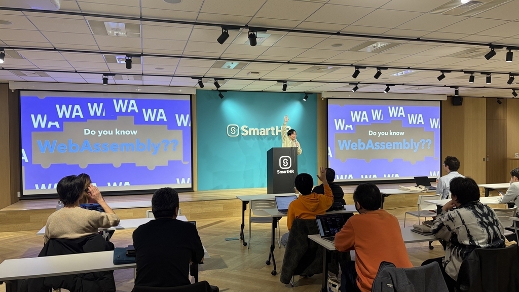 スクリーンに「Do you know WebAssembly?」と書かれたスライドが映っており、会場の参加者が挙手をしている