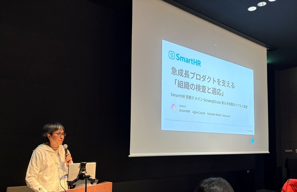 スポンサーセッションの発表直前のwassan。スクリーンには発表タイトルの「急成長プロダクトを支える「組織の検査と適応」&mdash;&mdash; SmartHR 労務ドメイン Scrum@Scale 導入半年間のリアルと展望」が映し出されている。