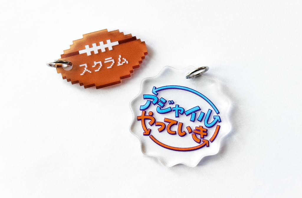 SmartHRの増えてくアクキーの新デザイン2つの写真。ドット絵のラグビーボールにスクラムと書かれた「スクラムラグビーボール」と、アジャイルの循環サイクルをイメージした円形の矢印がある「アジャイルやっていき」の2つのアクキー。