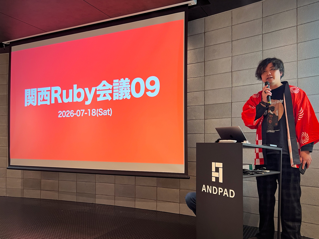 赤い画面のスクリーンの隣で赤いハッピを着た人がマイクを持って話している