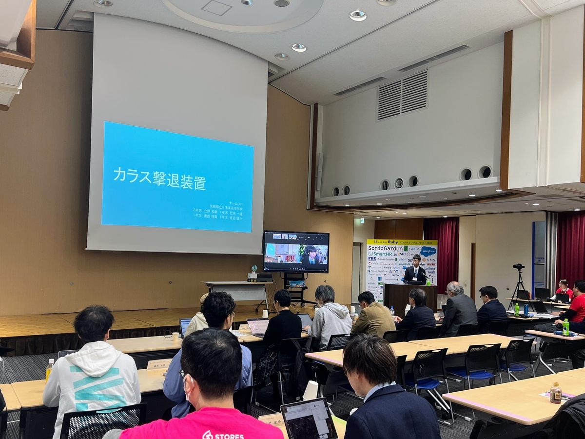 審査員や聴衆が見守るなか登壇している。壇上のモニターには、リモートから参加しているチームメンバーが映っている