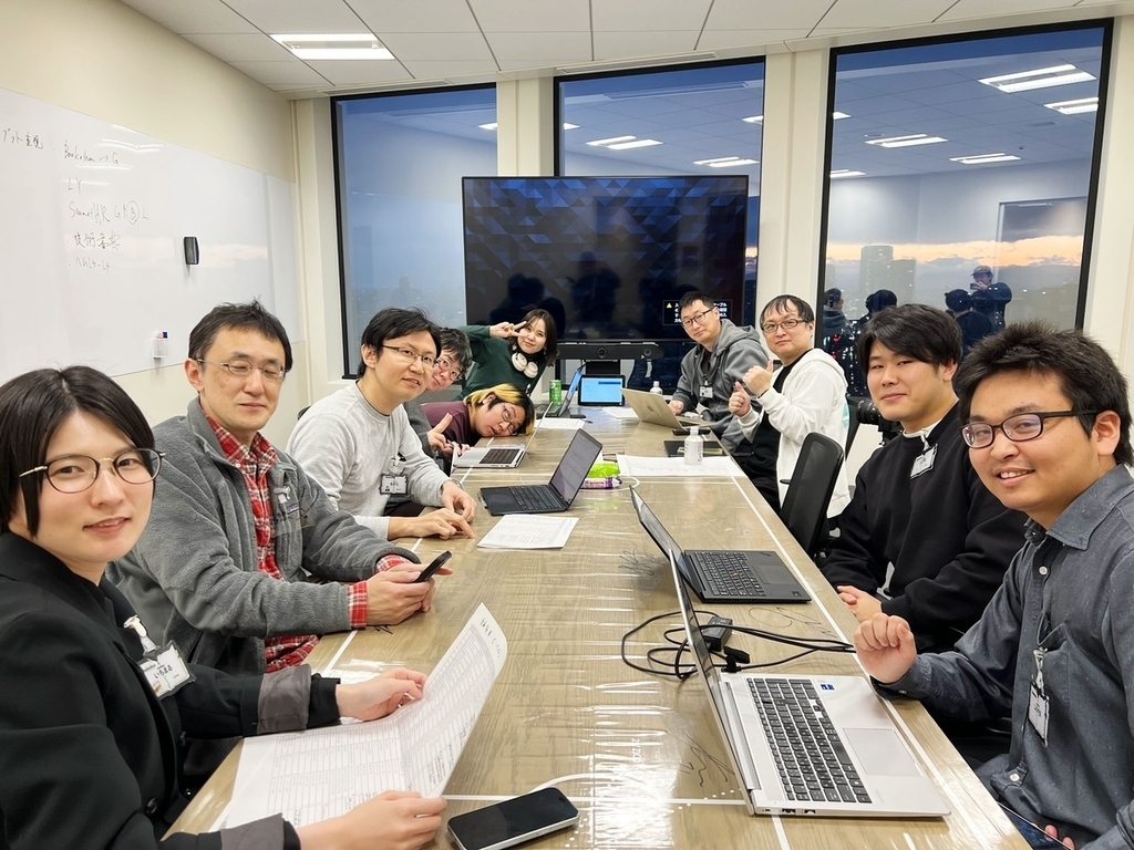 会議室のテーブルに座った10人のメンバーが笑顔でカメラを見ている