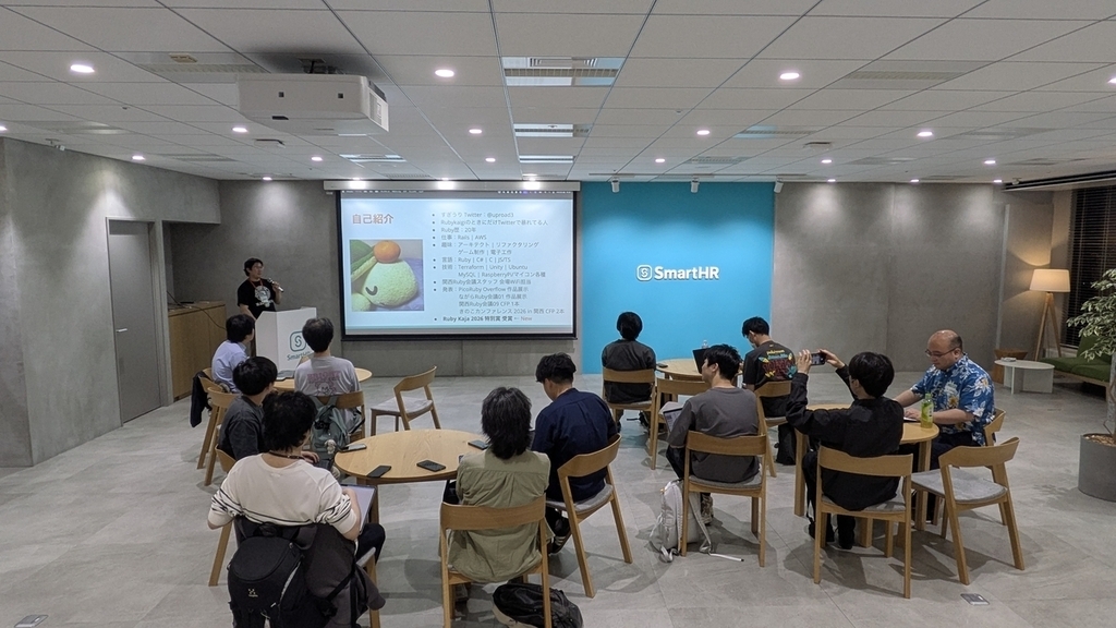 会場前方のスクリーン前で登壇者が話し、丸テーブルを囲んで着席した参加者がそれを見ている勉強会の会場全景