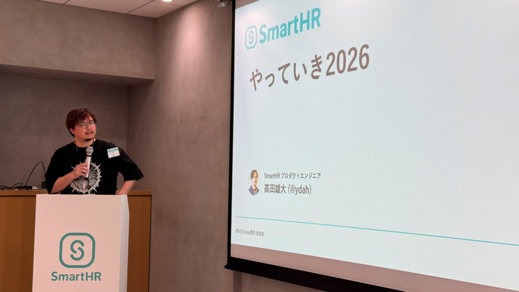 「やっていき2026」のタイトルスライドを背景に、演台の横でマイクを持って話す ydah