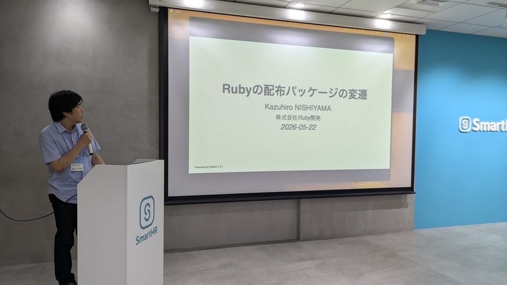 「Rubyの配布パッケージの変遷」のタイトルスライドを背景に、演台の横でマイクを持って話す znz さん