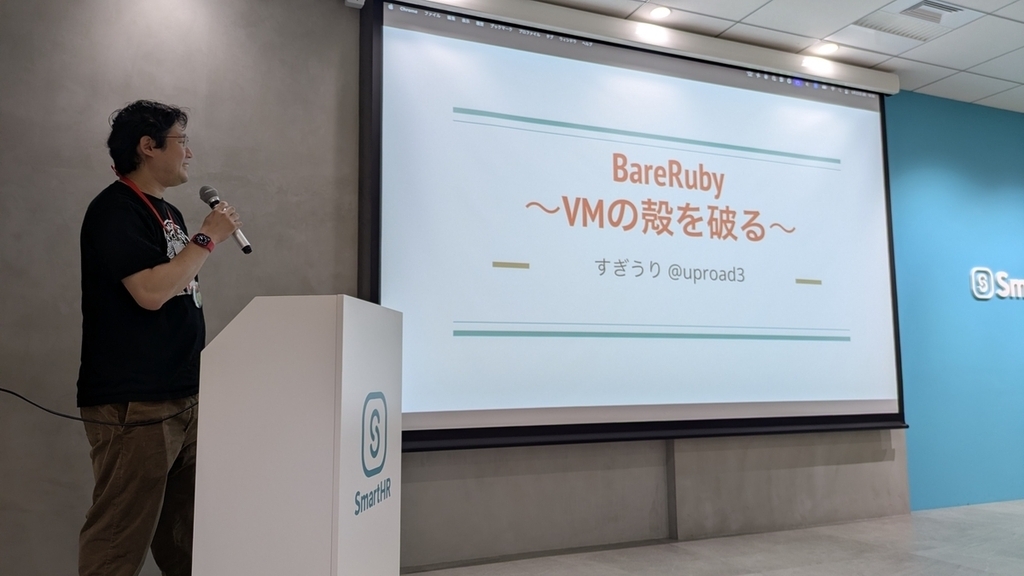 「BareRuby 〜VMの殻を破る〜」のタイトルスライドを背景に、演台の横でマイクを持って話す すぎうり さん