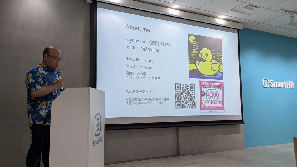 自己紹介スライドを背景に、演台の横でマイクを持って話す k-yoshida さん