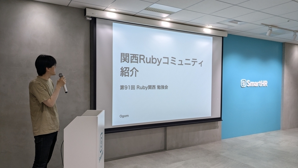 「関西Rubyコミュニティ紹介」のタイトルスライドを背景に、演台の横でマイクを持って話す おごもり さん
