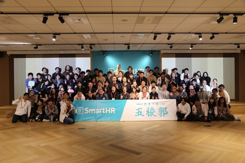 ステージ前に「SmartHR たまり場五稜郭」と書かれた白い横断幕を広げ、会場に集まったすべての参加者、登壇者、スタッフが笑顔で並んでピースサインなどをしている華やかな全員の集合写真