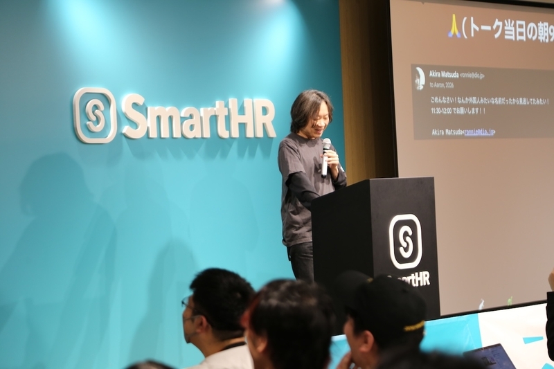 Aaron Patterson氏の自己紹介エピソードについて、チャット画面が写ったスライドを背に、マイクを持ってステージ中央に立ち、笑顔でRubyKaigiの温かい裏話を披露する松田 明さん