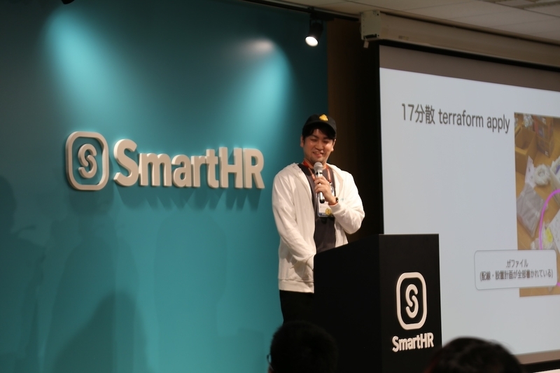 「17分散 terraform apply」という配線計画のスライドを映したスクリーンの横で、マイクを持ってNOCチームの奮闘劇をエネルギッシュに語るosyoyuさん