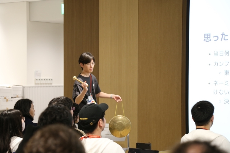 ステージ脇に佇み、大きな銅鑼（どら）を左手にぶら下げ、バチを右手に持って制限時間のタイマーをじっと見つめているterfnoさん