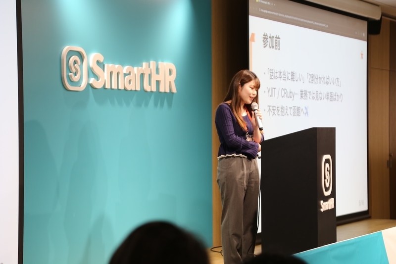 「初めてのRuby Kaigiはこう見えた」というスライドの前で、SmartHRの演台の後ろに立ち、両手でしっかりとマイクを握って客席へ熱く語りかけるasanoさん