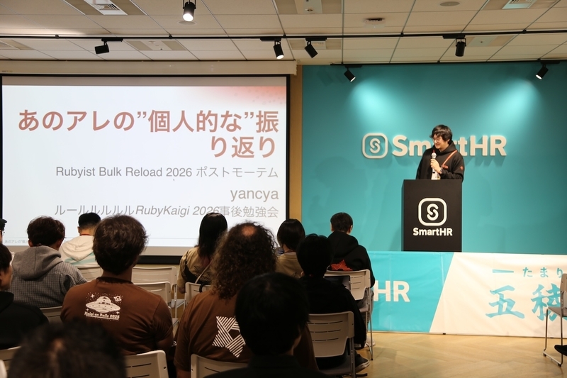 あのアレの”個人的な”振り返り」というスライドが大きく投影されたスクリーンの横で、SmartHRの黒い演台に立ちマイクを持って語るyancyaさん