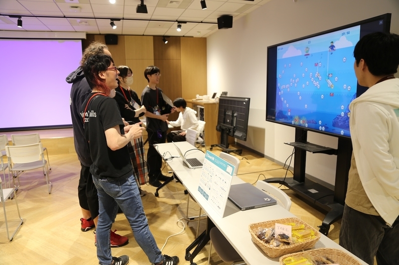 会場内に設置された大型液晶モニターに映るドット絵のイカ釣りゲーム画面を、専用の自作コントローラーを手に持ちながら楽しそうに見つめているエンジニアたちの後ろ姿