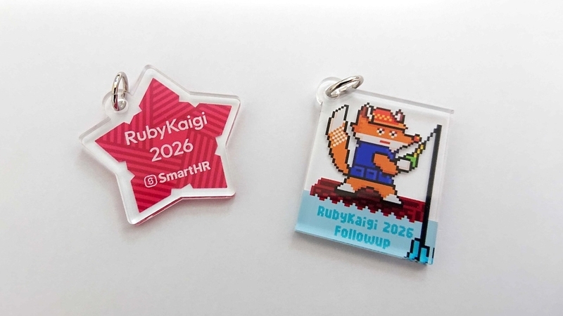 白いテーブルの上に並べられた2種類のアクリルキーホルダー。左側は星型に「RubyKaigi 2026」の文字とSmartHRのロゴが入り、右側はキツネがイカダに乗って釣り糸を垂らしているイラストに「RubyKaigi 2026 Followup」の文字がデザインされている。