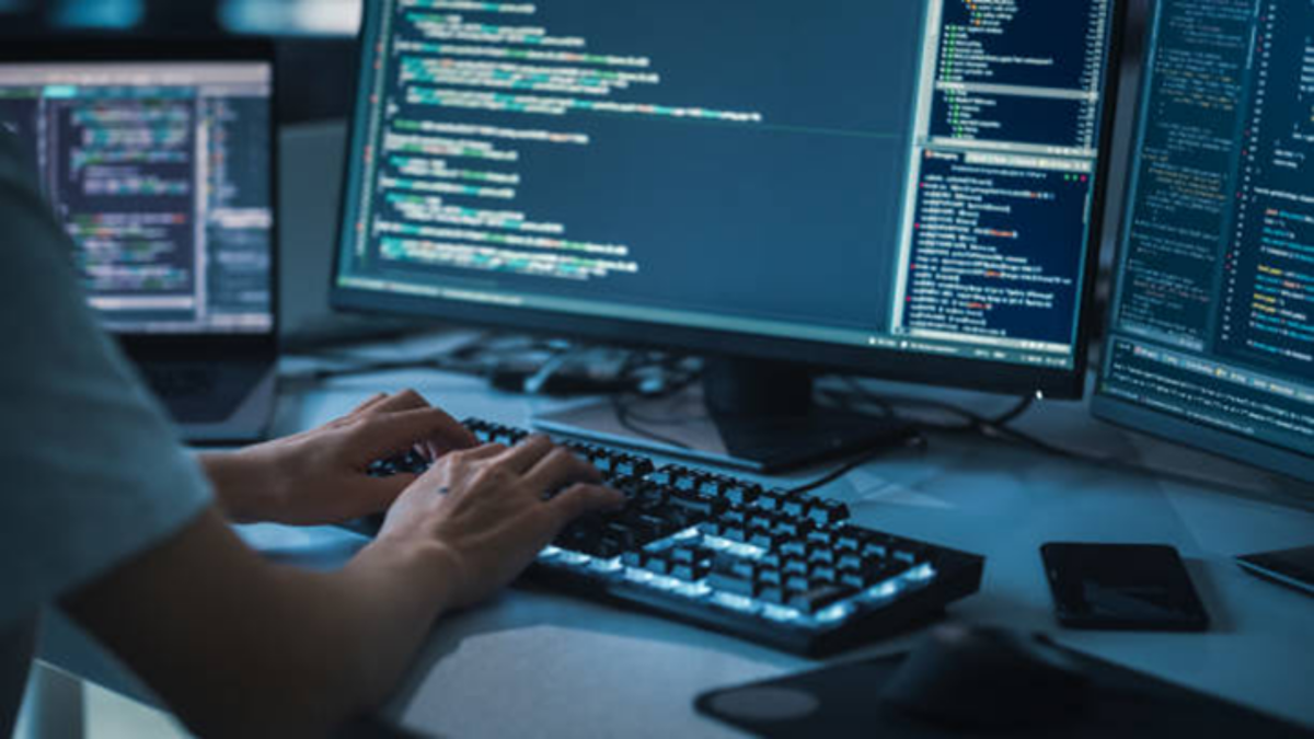 プログラミング用の複数モニターにコードが表示され、キーボードで作業するエンジニアの手元を写した写真。