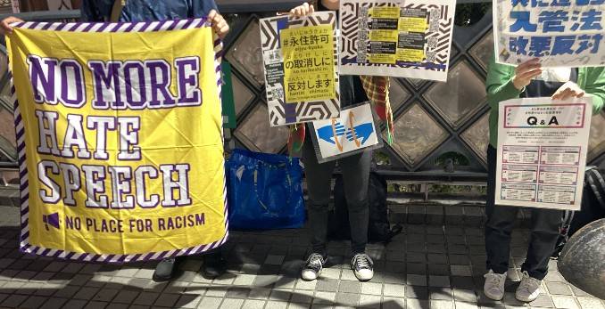 プラカードや風呂敷を持ってスタンディングをする3人の人の写真。風呂敷には「NO MORE HATE SPEECH NO PLACE FOR RACISM」と書かれており、プラカードは「#永住許可の取消しに反対します」「入管法改悪反対」と書かれている。また手話マークのプラカードを腰に巻いている人もいる。
