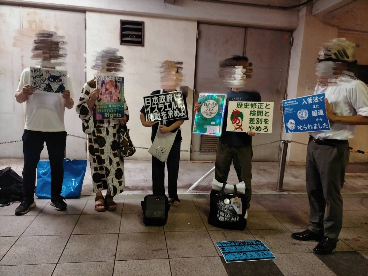 駅前で5人プラカードを持って立っている写真。プラカードは「入管法改悪反対」「入管法でまた国連から叱られました」「日本政府はイスラエルに停戦を求めよ」「歴史修正と検閲と差別をやめろ」というメッセージが並ぶ。他に難民・移民フェスの宣伝チラシも。宣伝チラシは電飾で装飾され様々な色で枠が光っている。