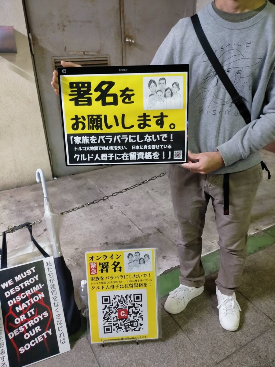 署名用プラカードを持つ人の写真。プラカードには「署名をお願いします。家族をバラバラにしないで！トルコ地震で住む家を住む家を失い、日本に身を寄せているクルド人母子に在留資格を！」と書かれている。地面にはオンライン署名のQRコードがつきプラカードが置かれている。