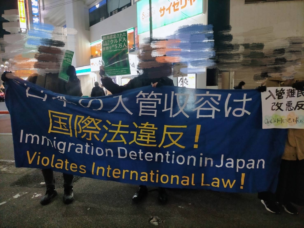 横断幕を持つ人々の写真。顔はモザイクがかけられている。横断幕には「日本の入管収容は国際法違反！Immigration Detention in Japan Violates International Law!」と書かれている。