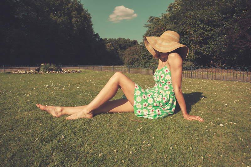 A young woman wearing a dress and a hat is sitting on the grass in a park