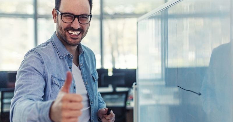 Young smiling modern developer standing in front of the whiteboard and giving thumb up