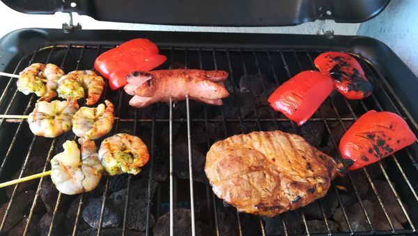 バーベキューのコンロの上の鉄板で、左手から串に刺したえび、セルブラ、肉、赤ピーマンが焼かれている。