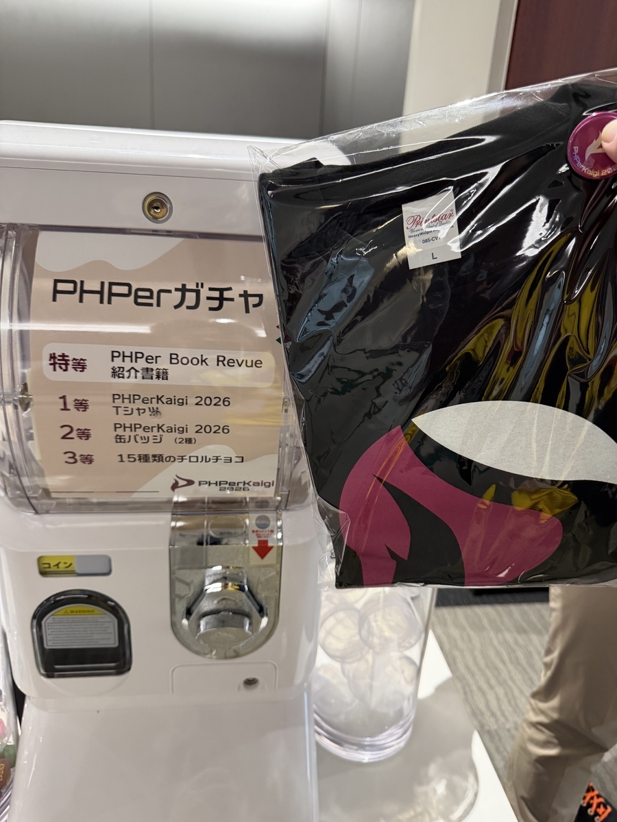 PHPerガチャのカプセルマシーンと、ビニールに包まれた黒いPHPerKaigi Tシャツ、小さい缶バッジ