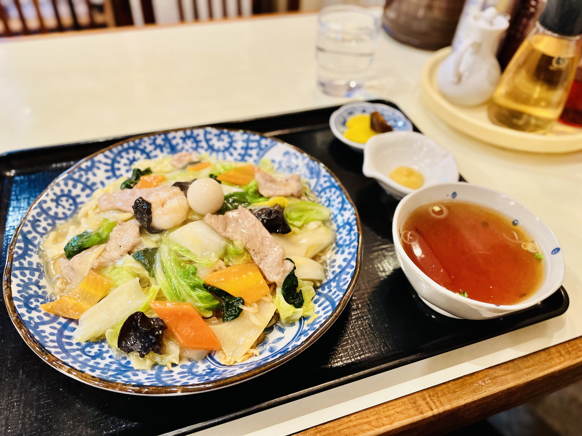 大皿に平たく盛られたあんかけ焼きそば。たくさんの野菜と、きくらげ、大きなエビ、うずらの卵などが乗っている。シンプルなスープと漬物付き。絵を小皿に入った和からしもあり、これをお酢で溶いてかける。