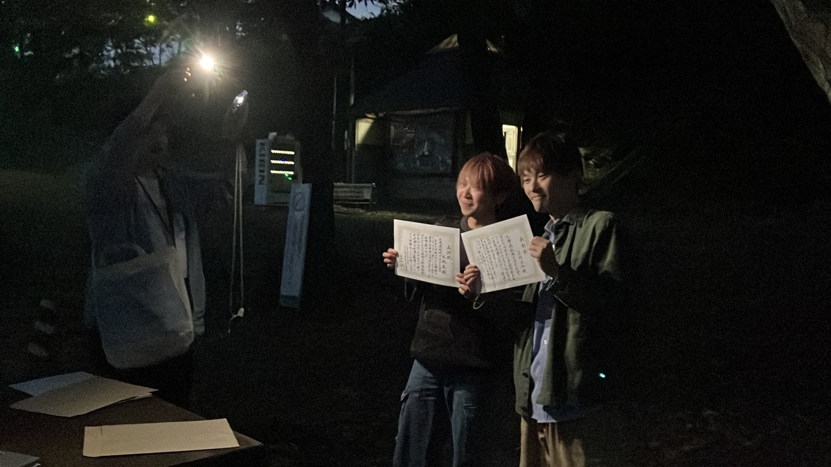 暗い公園の中でスマートフォンの灯りに照らされた 0yu くんとやまとさんがそれぞれ授与を受けた賞状を持っている様子