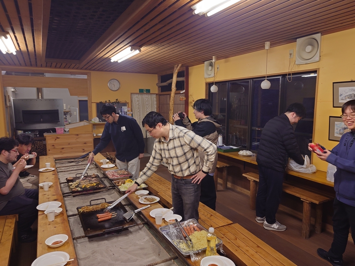 屋内テーブル中央に鉄板を２枚置いてバーベキューを楽しむ８人の参加者