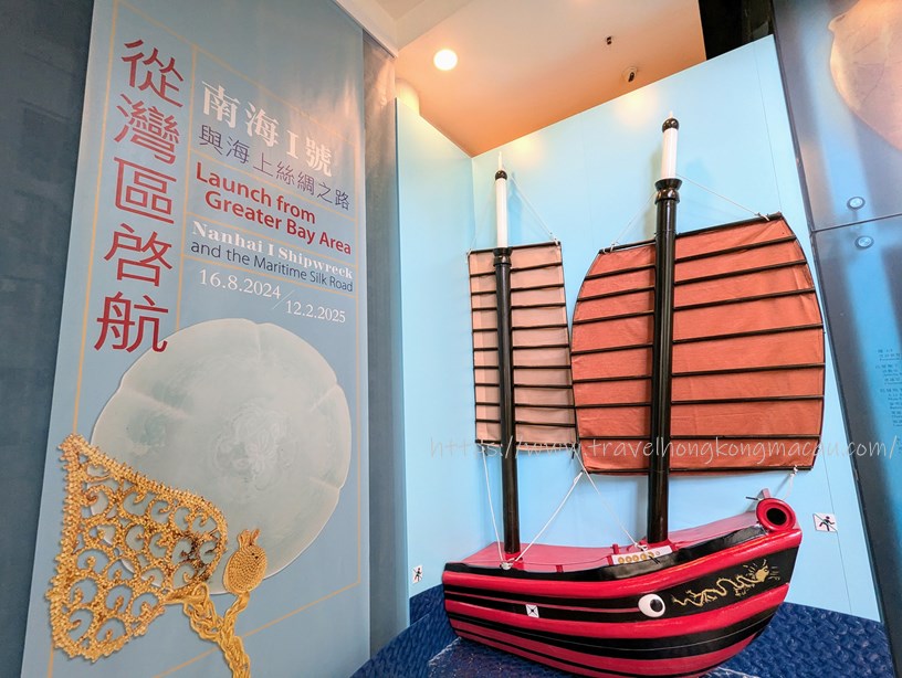 A traditional Tang Dynasty-style wooden boat with red sails displayed against a light blue background, next to a poster about the launch from Nanhai I Shipwreck and the Maritime Silk Road.