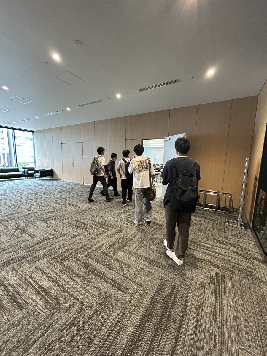 グレーの絨毯がひかれた室内の写真。奥には壁と扉があり、扉に向かって5人の人間が歩いている様子。