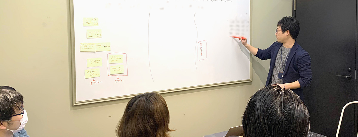写真：ホワイトボードを書きながら説明する人と、聞き入る人