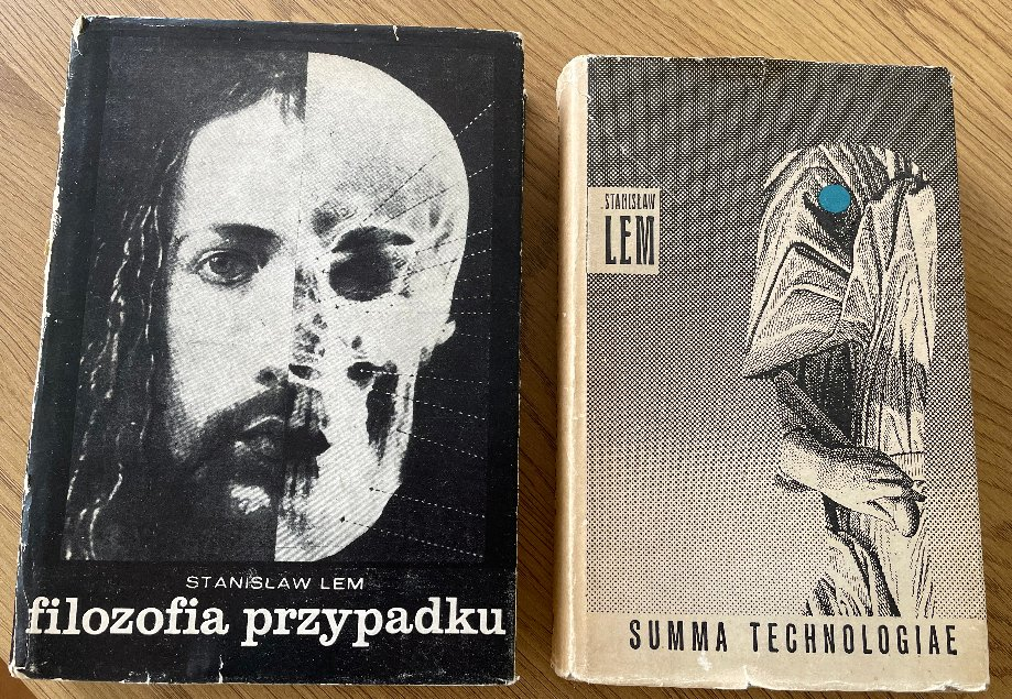 "木のテーブルに置かれたスタニスワフ・レムの2冊の古書。左側は『filozofia przypadku』（偶然の哲学）初版で、表紙は左半分が髭を生やした男性、右半分が髑髏の白黒デザイン。右側は『SUMMA TECHNOLOGIAE』（技術大全）で、青い点のあるローブ姿の人物イラストにも見える抽象画が黒の点描で描かれている"