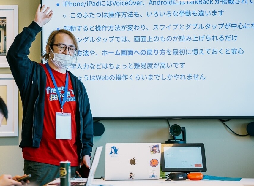 講師（筆者）が手を挙げ、挙手を促している。もう片方の手にはスマートフォンを持っている。赤い「技術の日」Tシャツのうえにパーカーを羽織っている
